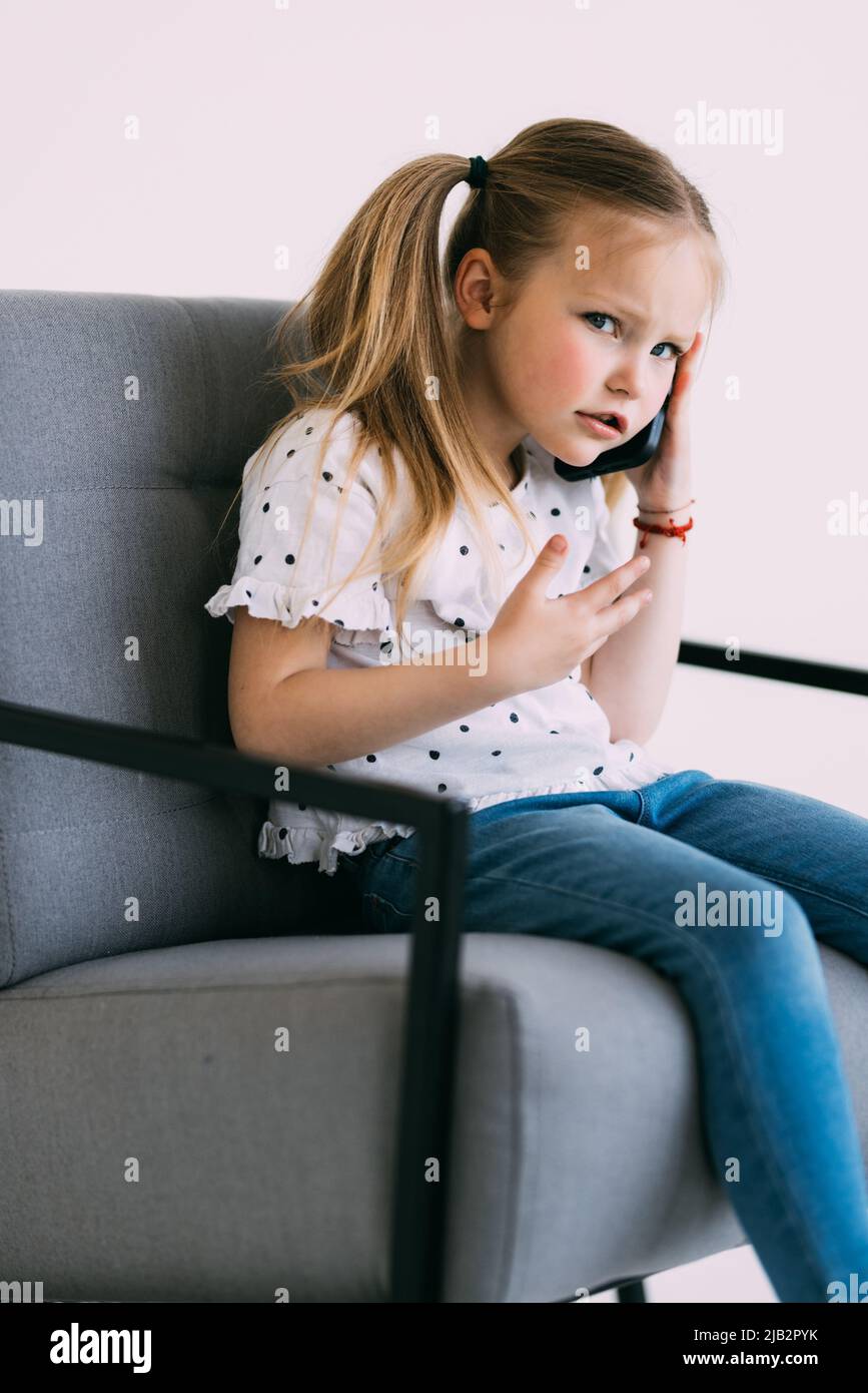 Young Little Confused Girl Talk Phone Sit On Chair On White Background young-little-confused-girl-talk-phone-sit-on-chair-on-white-background