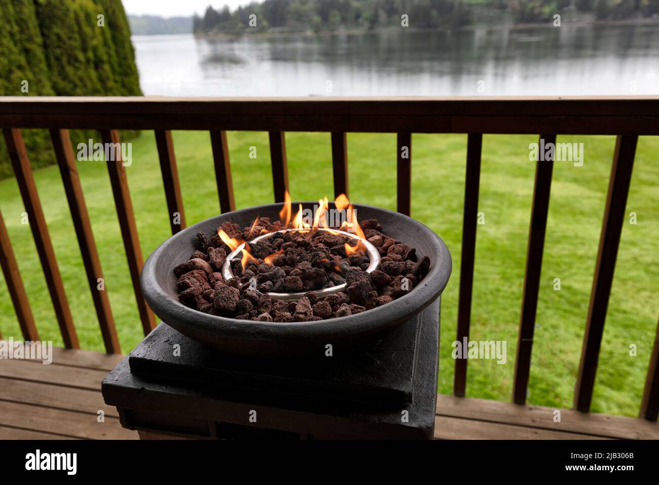 Burning gas fire pit, natural lava stones, on outdoor home deck with lake and woods in the background Stock Photo