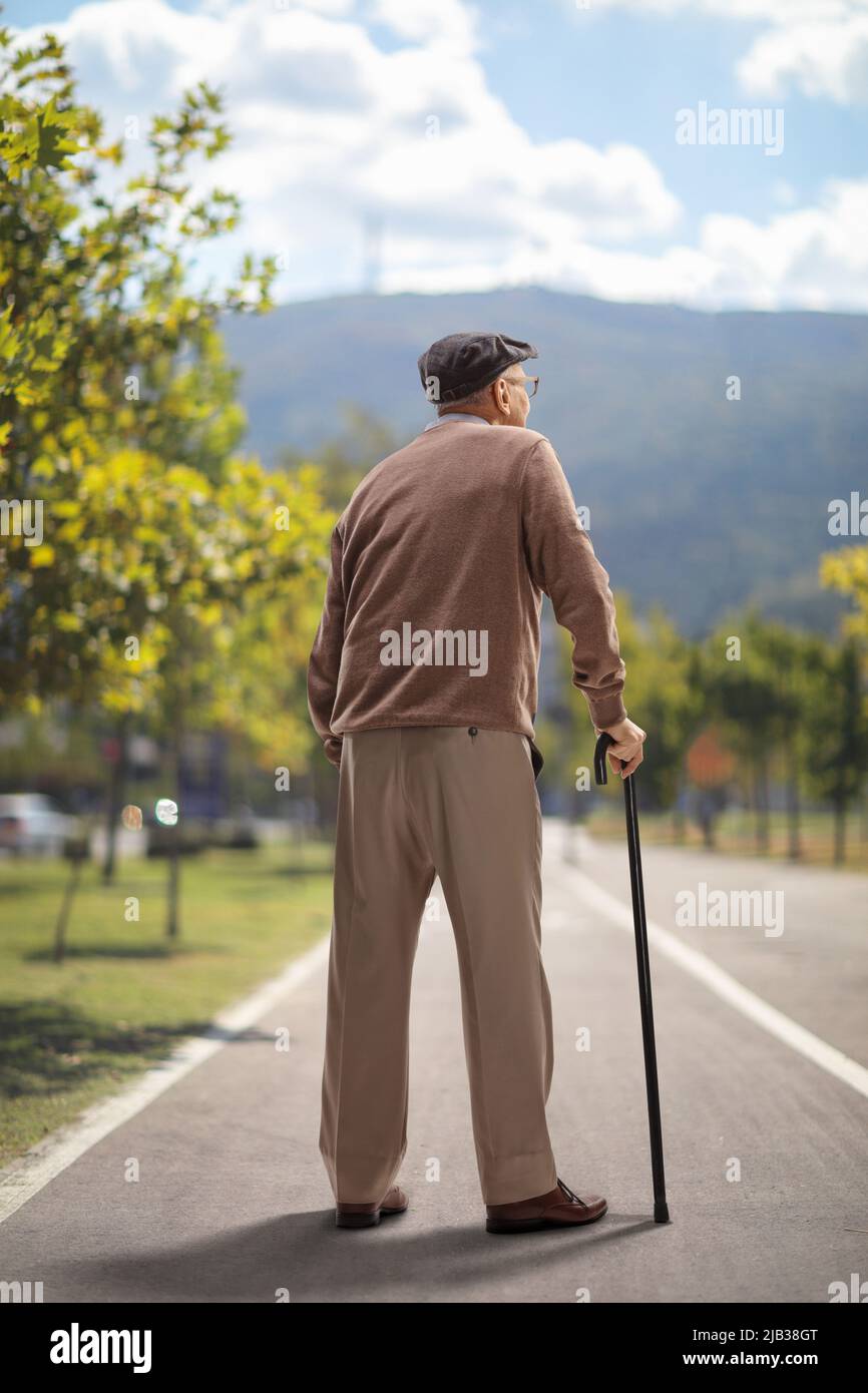 Rear view shot of an elderly man standing with a cane on an asphalt ...