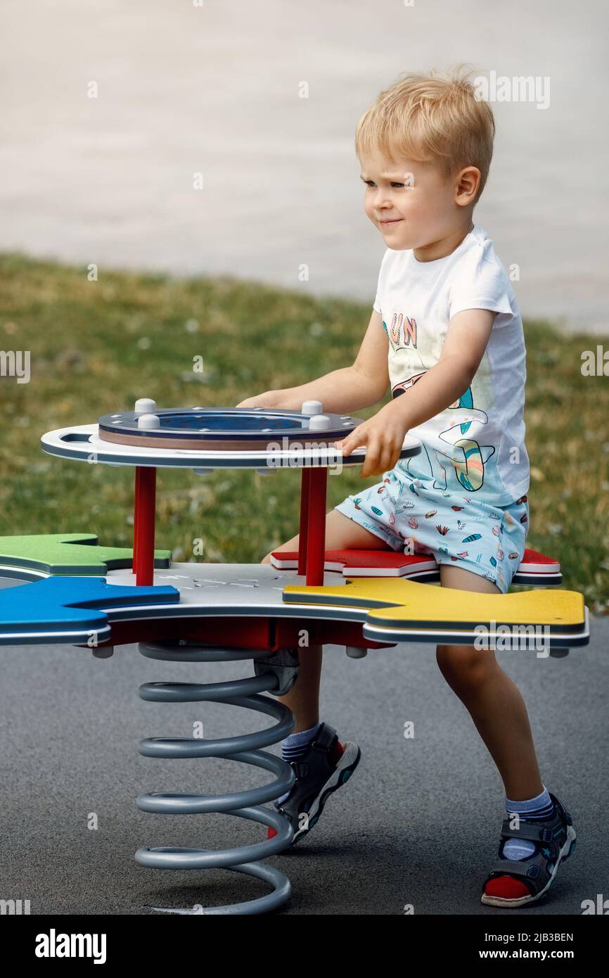 The cheerful boy springs up and balances on the swings of a modern outdoor playground. Stock Photo
