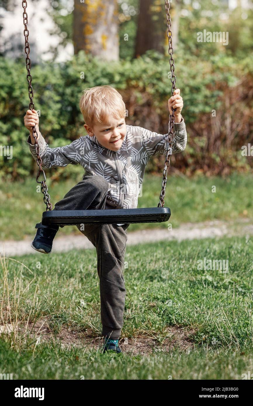 The blond boy plays with a chain swing as he tries to climb on it without his parents help. Stock Photo