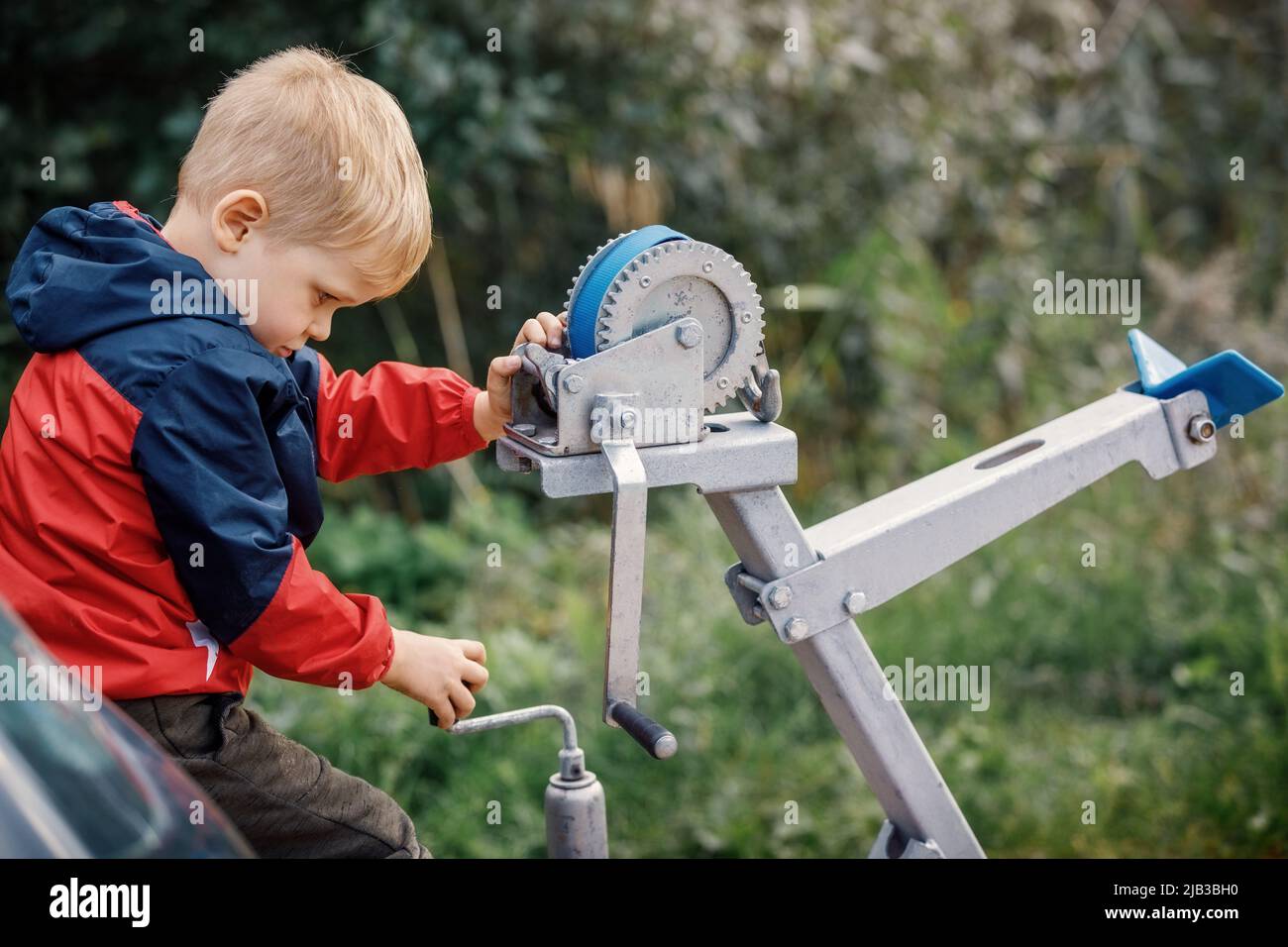 The little boy concentrated and playing with boat trailer winch, boat bow rest, blue cargo strap and its pulley and gears. The child turns the knob. Stock Photo