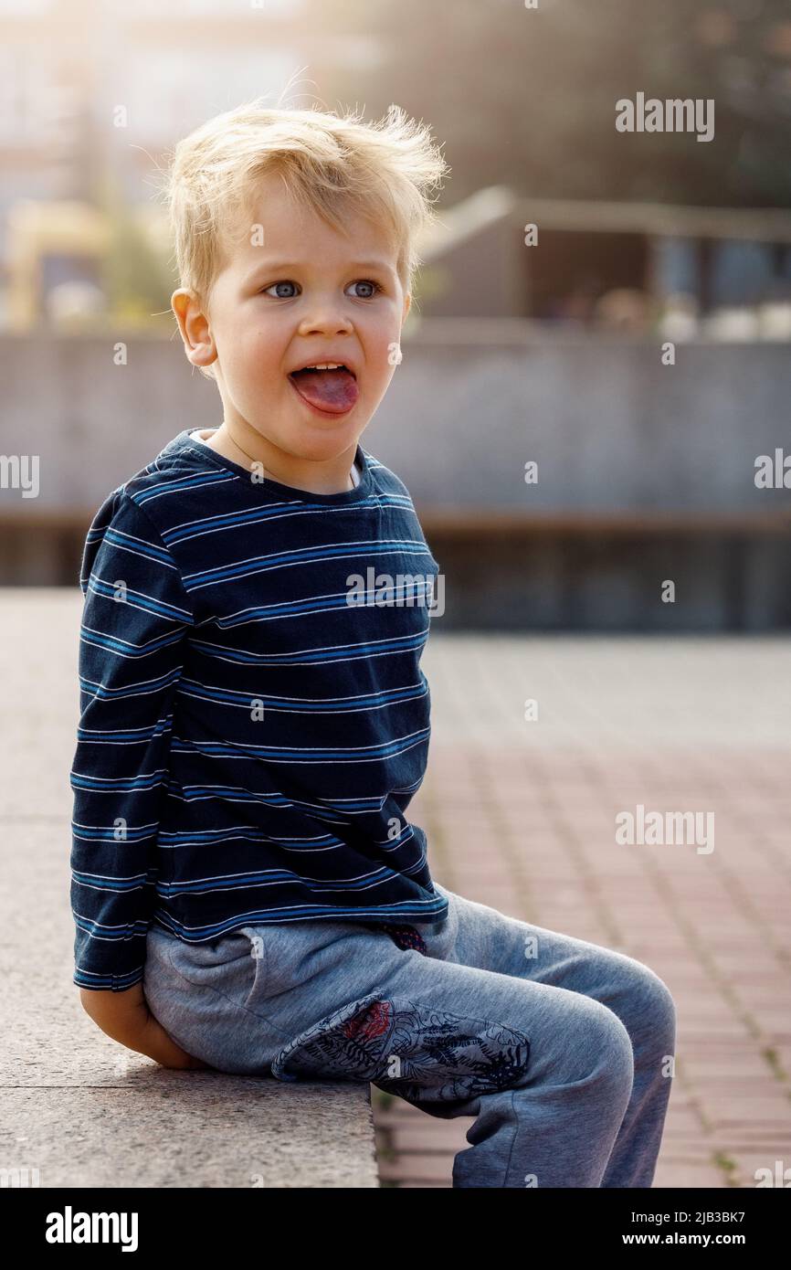 A portrait of a little naughty, sympathetic boy sitting by a city fountain, the child poses, showing his tongue. Stock Photo