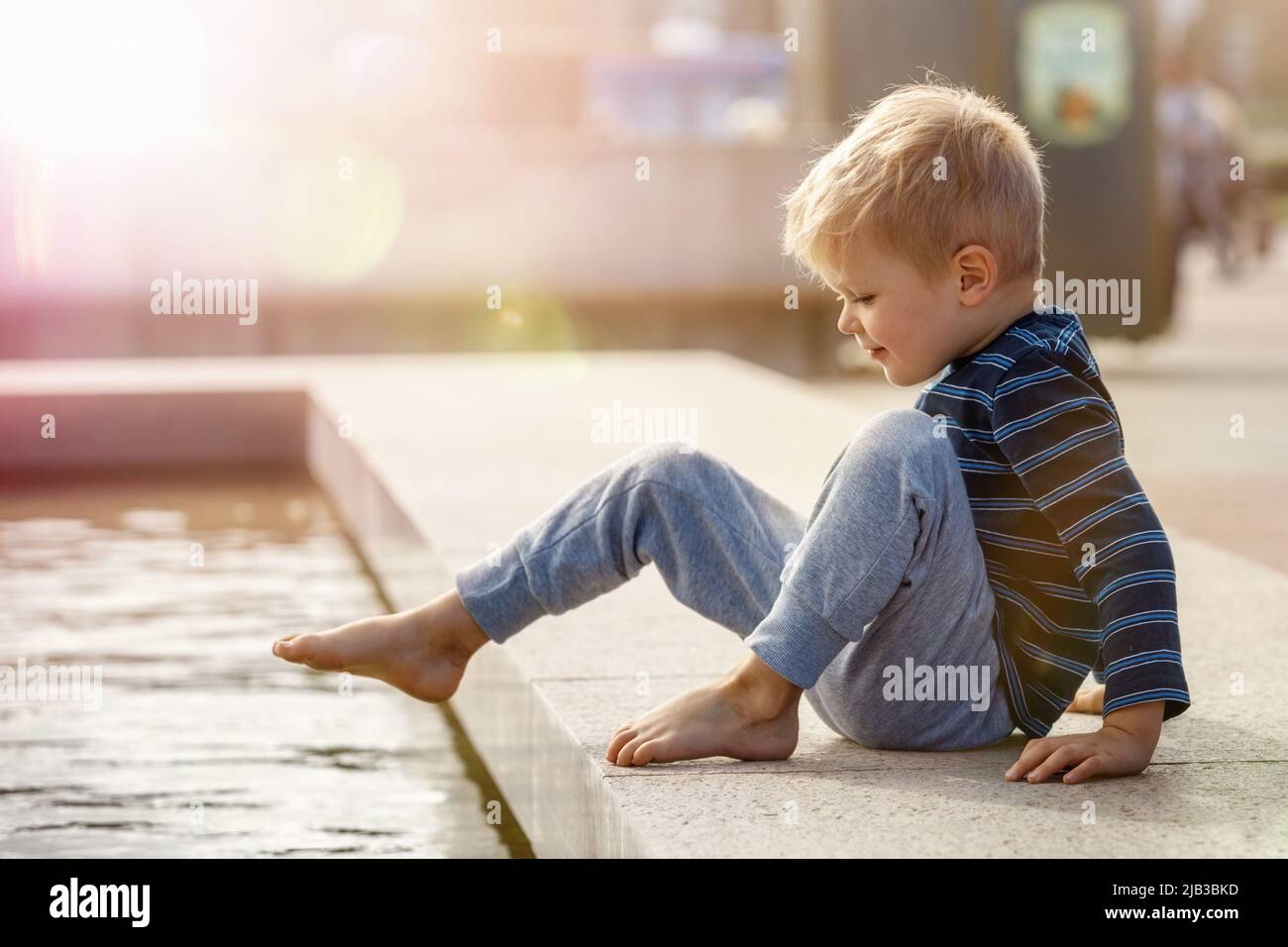 Cute little boy playing with water hot summer outdoors, the child tests the temperature of the water in the fountain by putting his foot into the wate Stock Photo