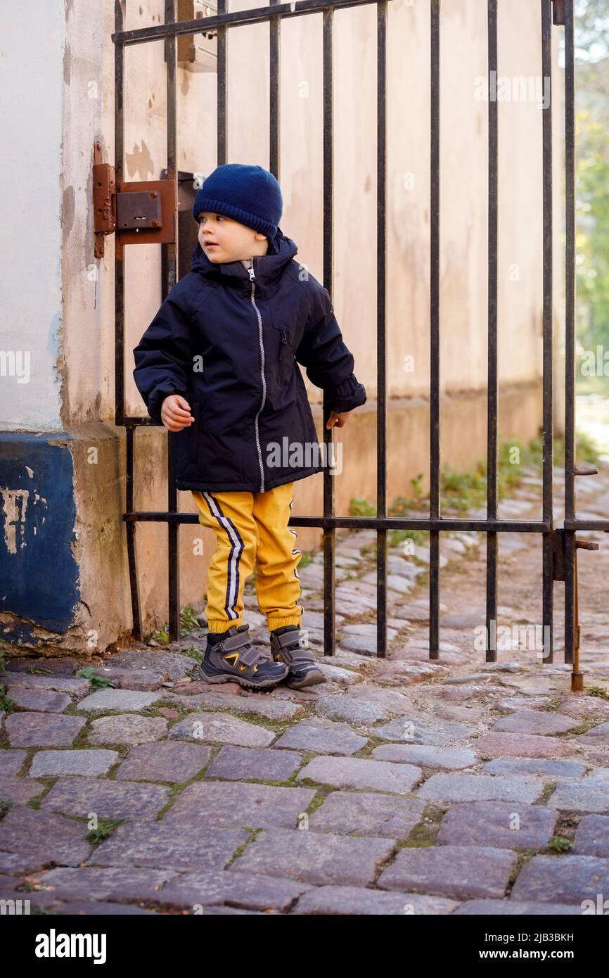 Little careful boy in in the old town. Lovely kid looking back to make sure no one sees him, he tries to get through the gate into the yard. Outdoor w Stock Photo