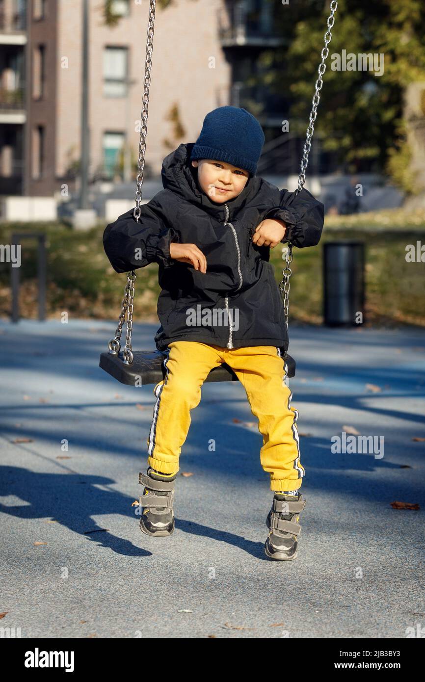 A cute boy in warm autumn clothes, yellow pants and a blue hat swings on a chain swing with a smile. Stock Photo