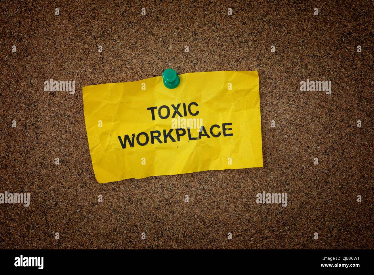 A crumpled sticky note with the words Toxic Workplace on it pinned to a cork board. Close up. Stock Photo