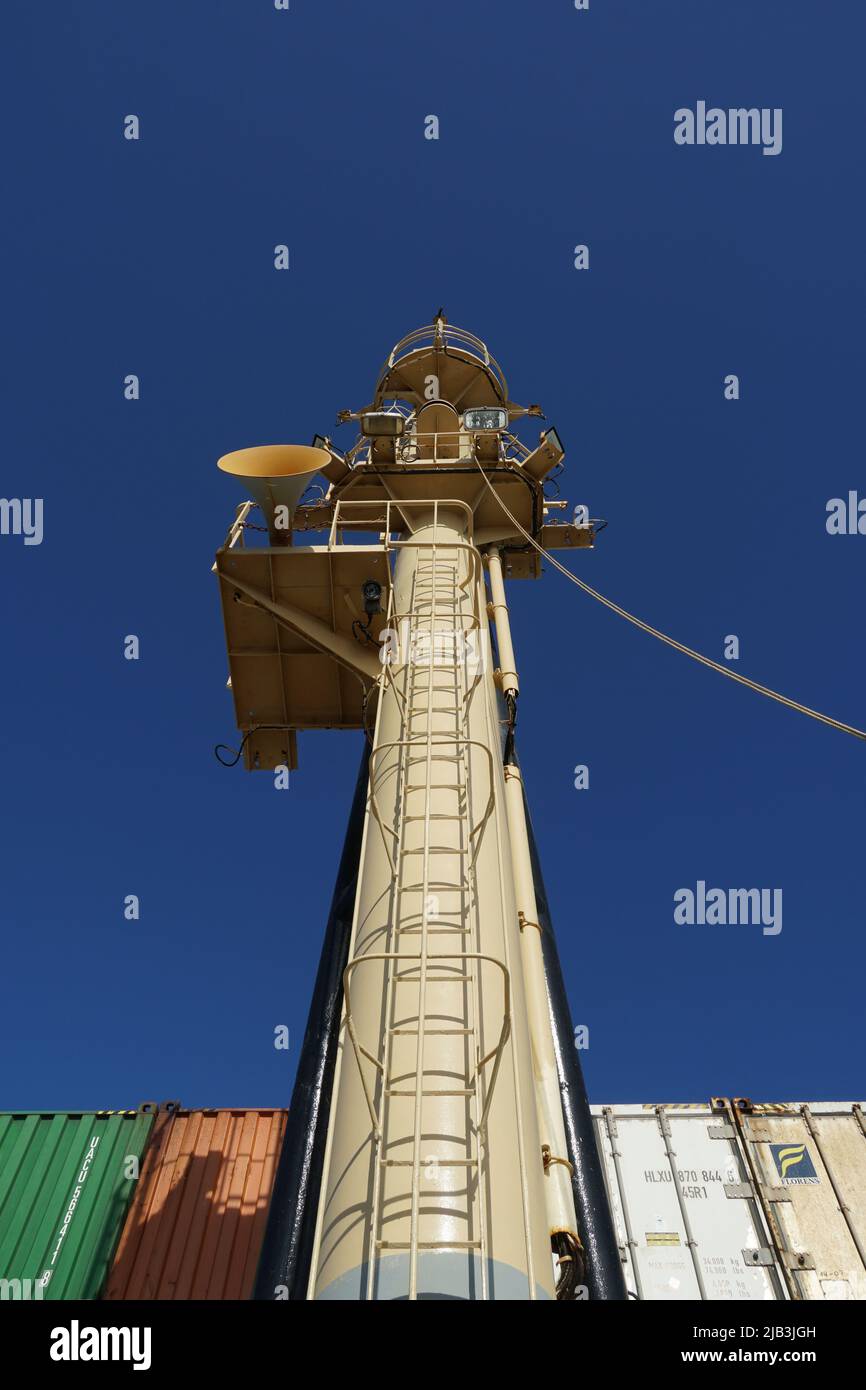 View on forward vessel mast with ladder and foghorn from mooring station, from deck to top.  Behind are stowed containers from different shippers. Stock Photo