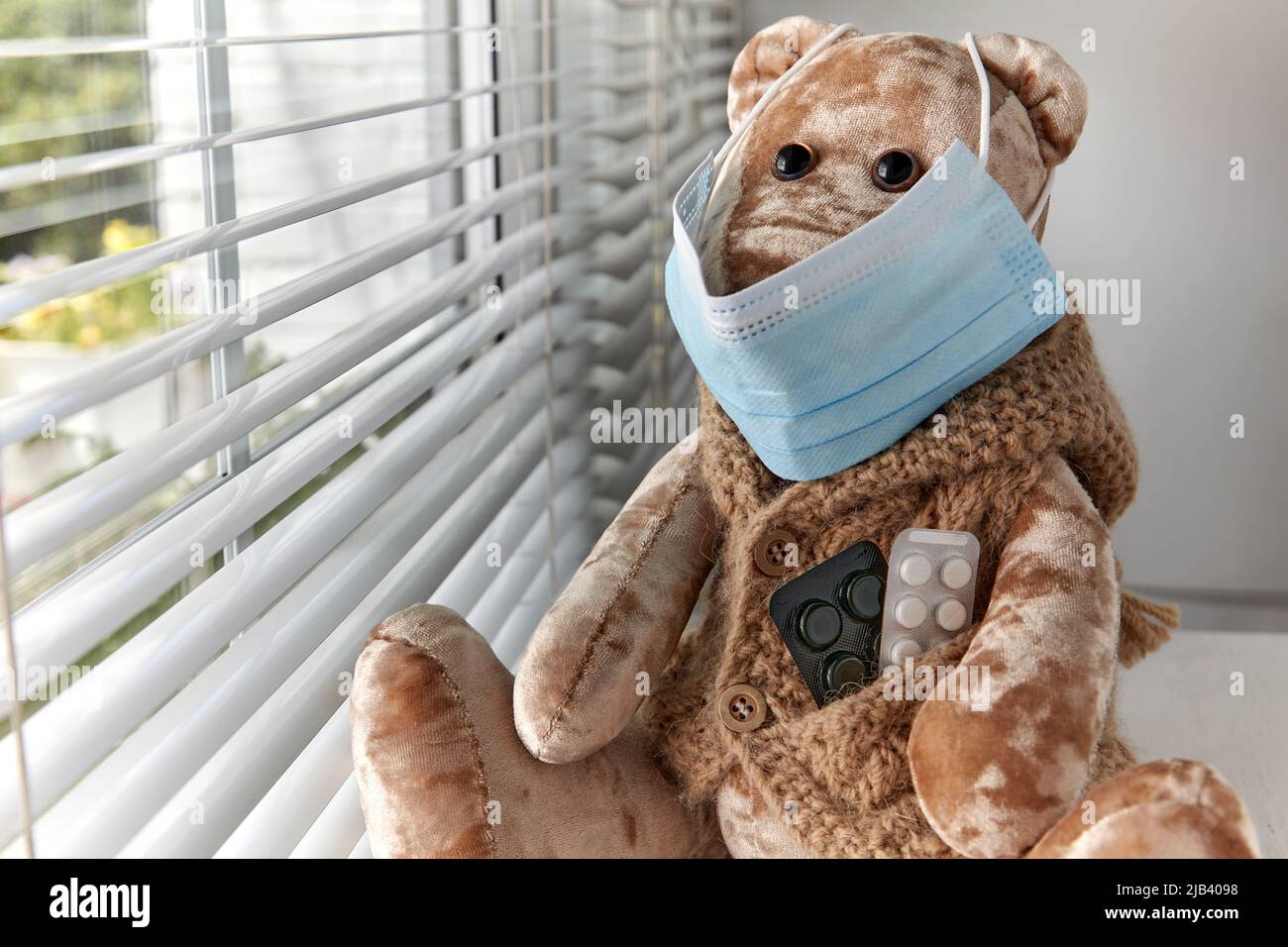 A teddy bear in a medical mask looks sadly out the window. Sick child ...
