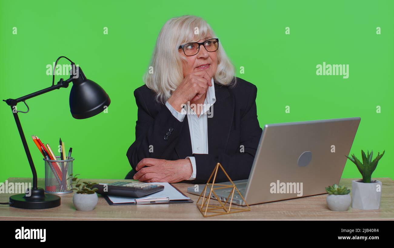 Senior businesswoman in thought and conjectures looking to side, dreaming, imagining something pleasant fantasizing about weekends holidays. Elderly woman isolated on green chroma key studio at office Stock Photo