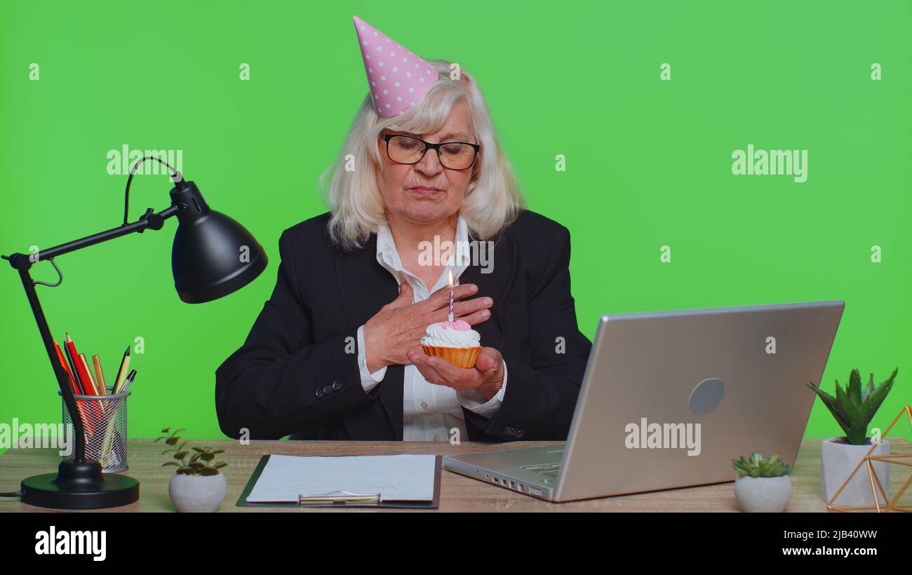 Sad upset lonely senior businesswoman in festive hat celebrating birthday party alone, feel frustrated despair disappointed sadness holds cake with candle at office. Elderly woman on green chroma key Stock Photo