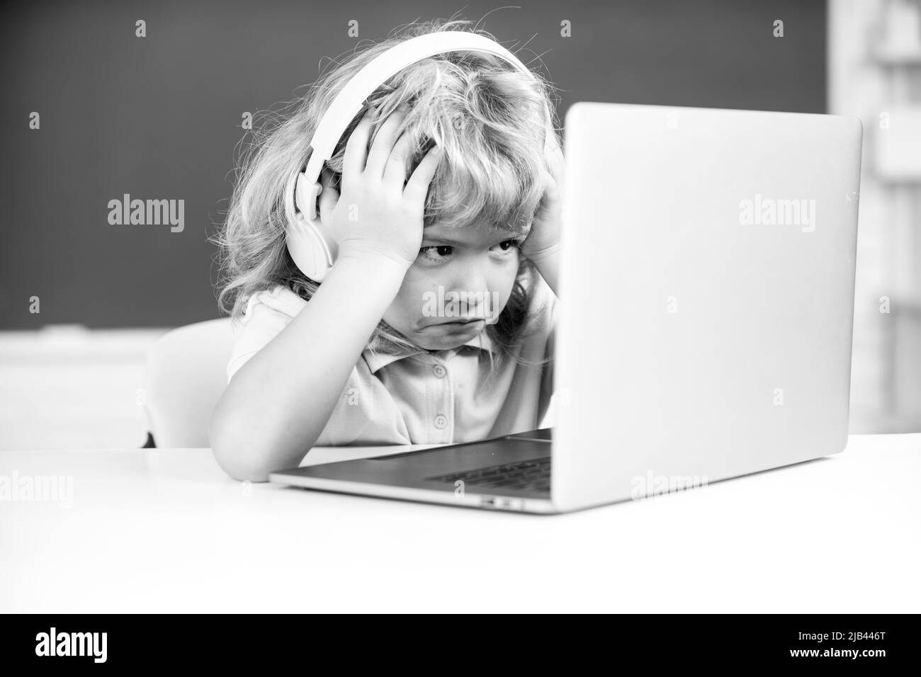 Angry sad boy in headphones sit at desk, study online on laptop at school, klever kid wear earphones in notebook learning using internet lessons. Stock Photo