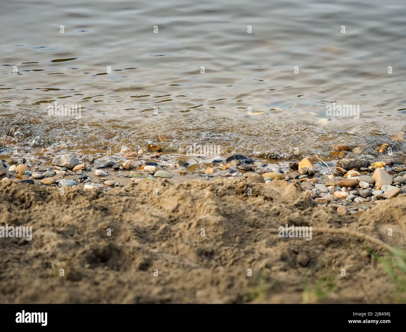 Water waves are moving to the shoreline of a river or a lake. Natural motion and fluidity. Wet stones and sand on the coast. Close-up of a beach. Stock Photo