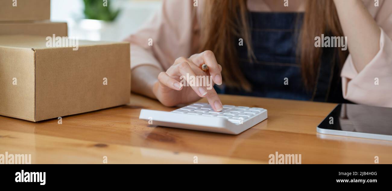 Asian woman entrepreneur using calculator with pencil in her hand, calculating financial expense at home office,online market packing box delivery Stock Photo