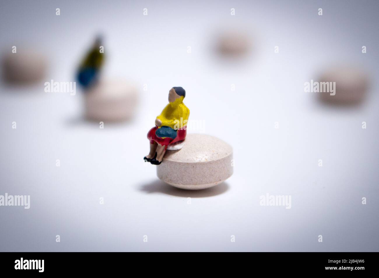 Miniature old woman in yellow outfit sitting on a pill in the white bg. Tablets are randomly placed and the woman keeps distance away from the guy. Stock Photo