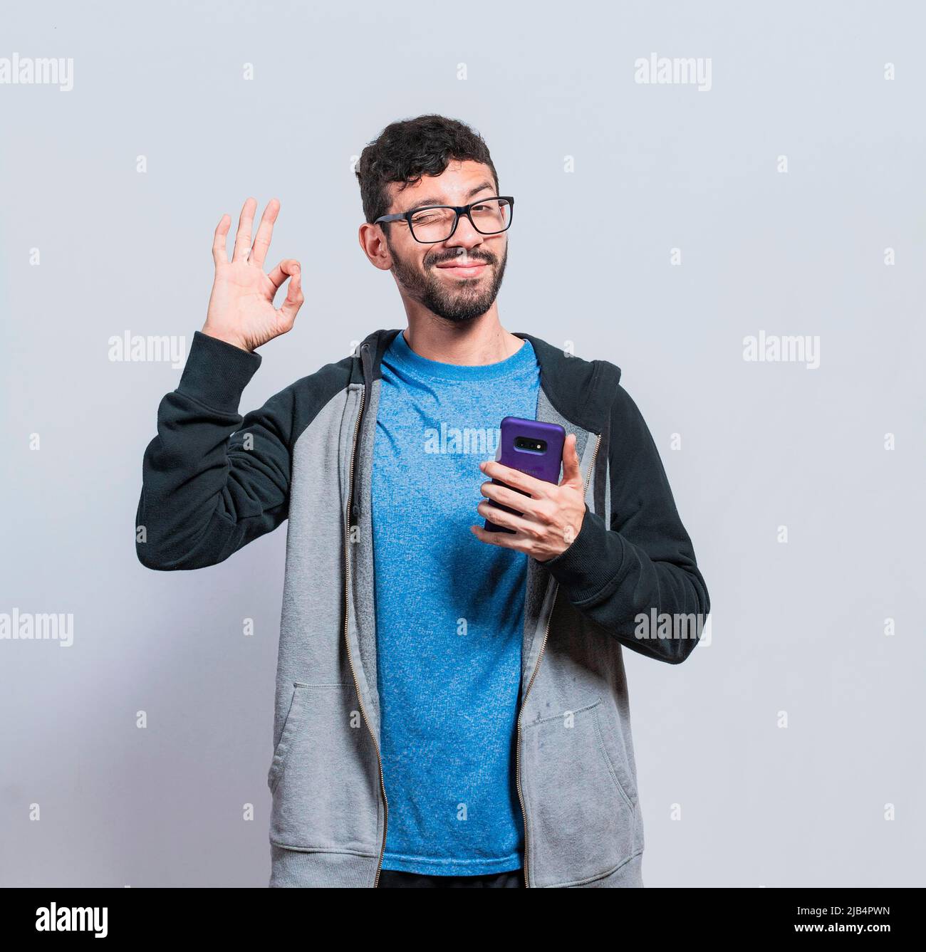 Pleasant person holding cellphone and winking, smiling boy with cellphone making ok sign, concept of positive person with cellphone closing fingers Stock Photo