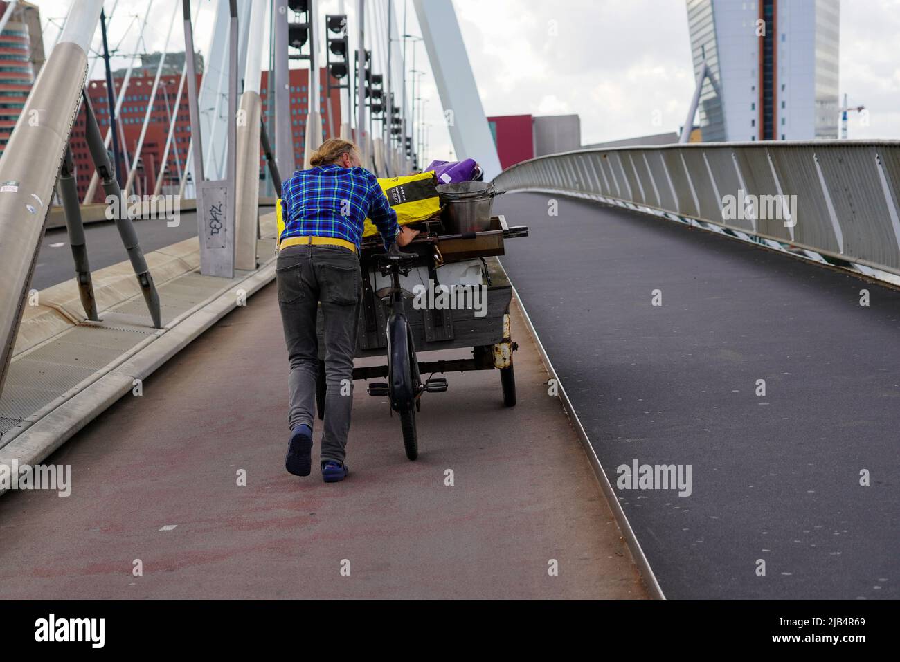 A man pushes his cargo bike  in windy weather across the Erasmus Bridge in Rotterdam, Netherlands, 5/27/22 Stock Photo
