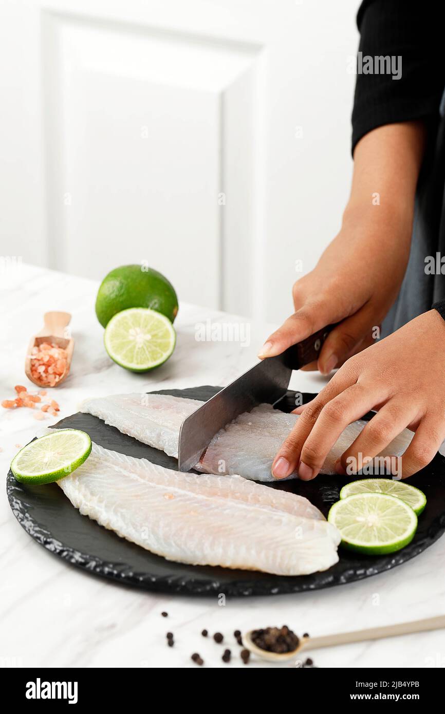 Female Hand Cutting Dori Fillet Pangasius Fish above Stone Board, Process Cooking in the Kitchen Stock Photo