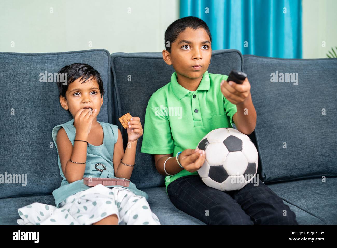 sibling kids busy watching television or tv by sharing biscuits at home - concept of holidays, caring and leisure activities Stock Photo