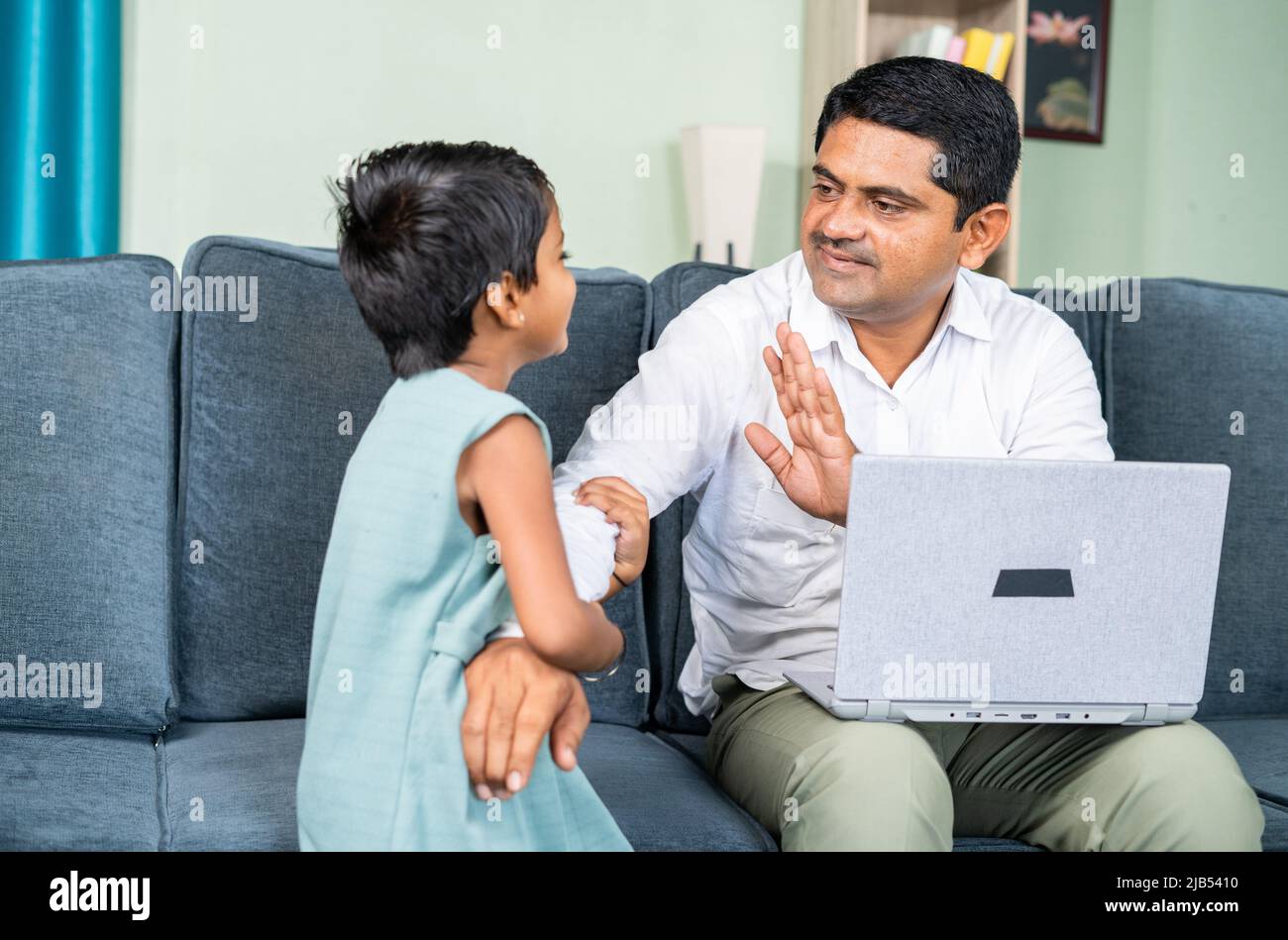 little daughter disturbing by pullinf her father while busy working on laptop at home during coronavirus covid-19 pandemic - concept of work from home Stock Photo