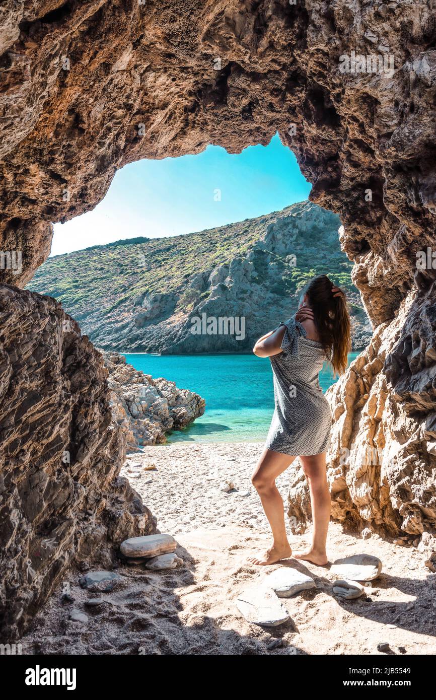 A young girl in a gray dress with long hair stands in a cave by the sea.  View of the paradise beach on the Aegean coast of Greece. Cave in the sea