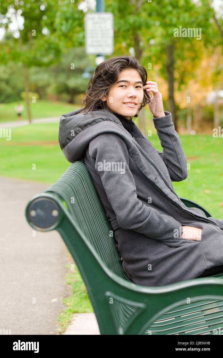 Smiling biracial teen girl or young adult female in gray jacket sitting on green park bench by lake on overcast autumn or spring day Stock Photo
