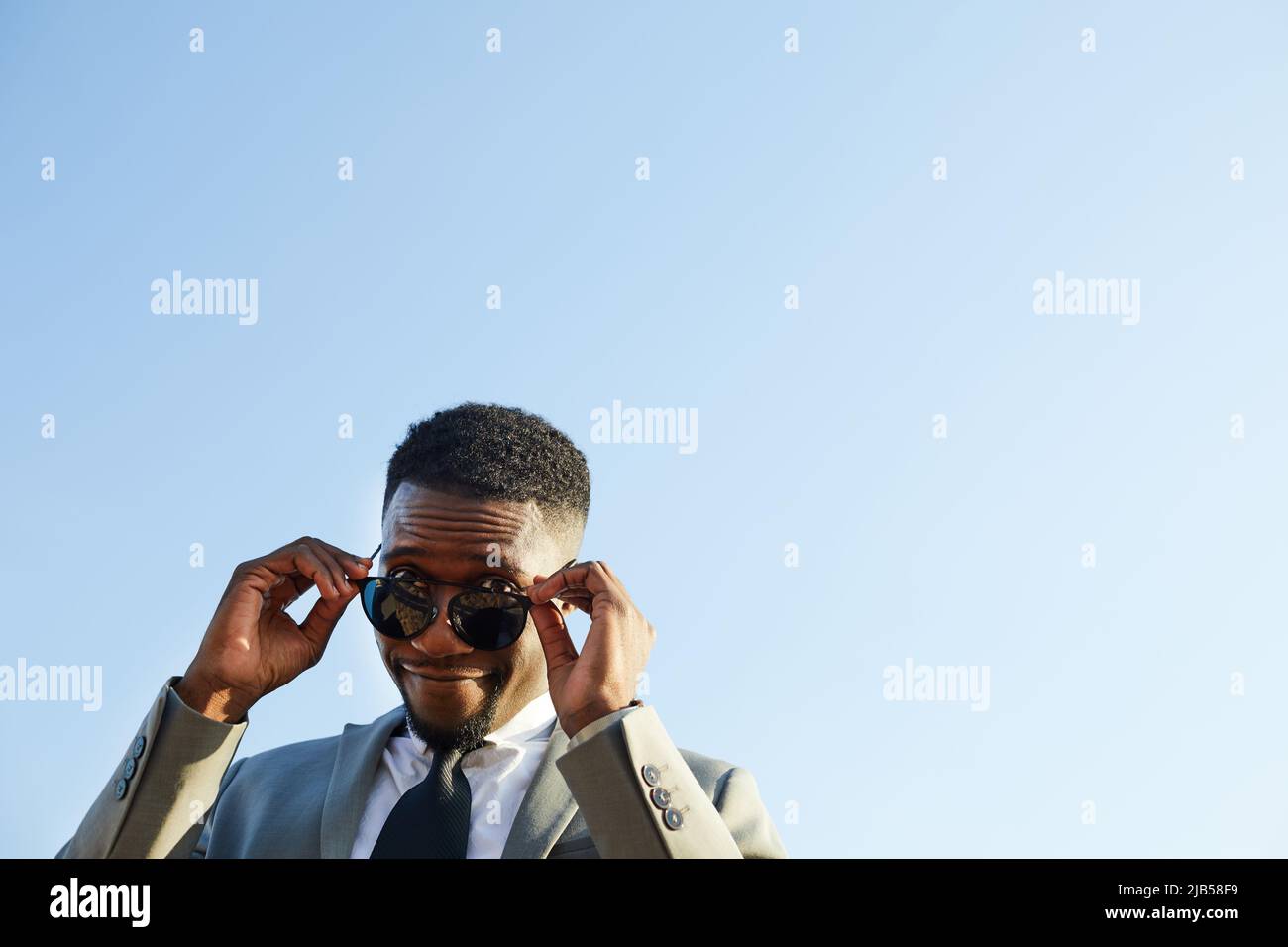 Content frowning smug young black businessman in suit adjusting sunglasses and looking at camera while standing against sky Stock Photo