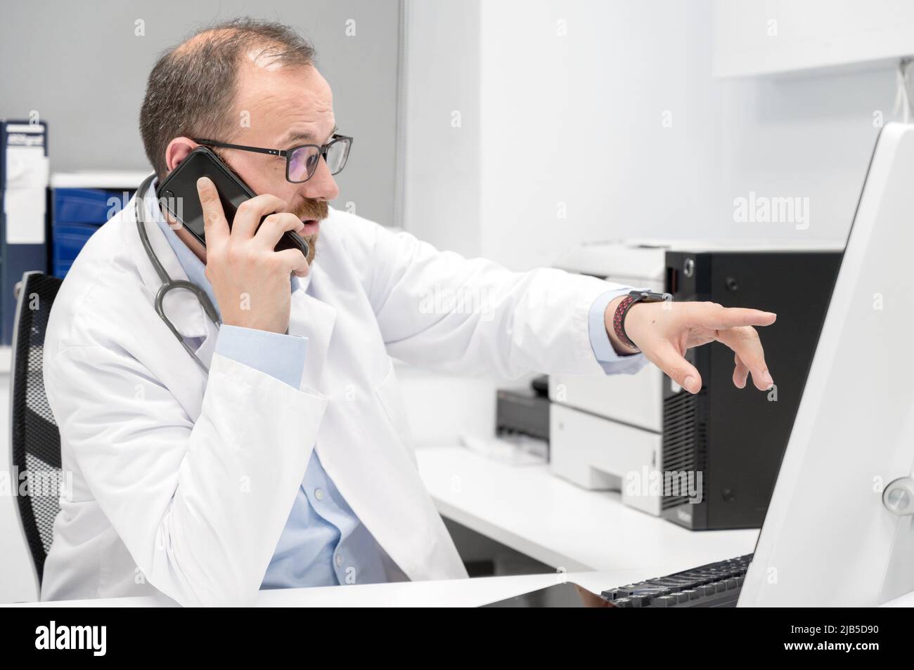 Male doctor pointing with finger on desktop computer while talking on the phone, discussing treatment with colleague. High quality photo. Stock Photo