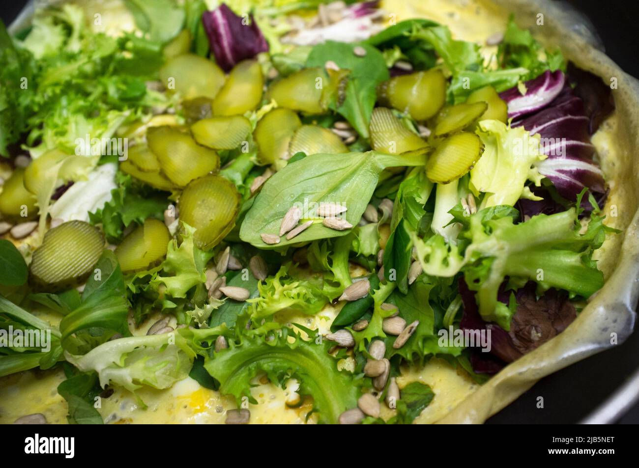 Hot salad over  scrambled egg on pan. Plate with several lettuces, gherkins and sunflowers seeds. Stock Photo
