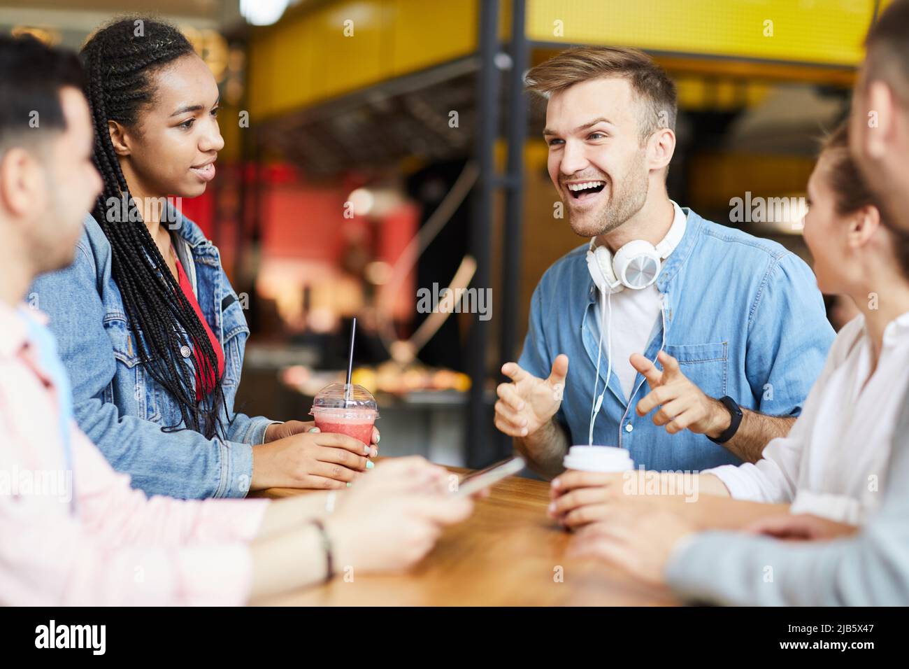 Emotional handsome hipster man with headphones on neck sitting at table and telling story to friends in cafe Stock Photo