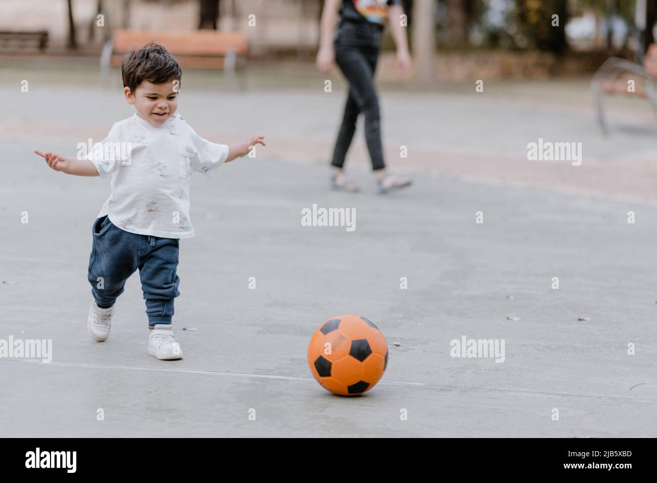 Little boy playing with a soccer ball in the park, his mother watching him for safety Stock Photo