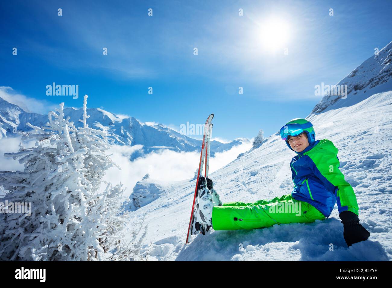 Preteen boy with alpine ski sit in snow over mountain peaks Stock Photo