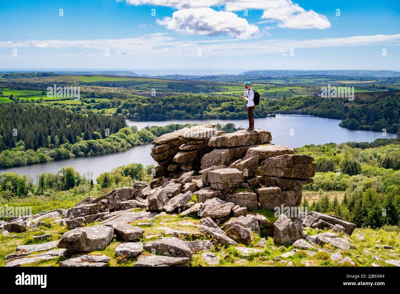 A walker stands on a granite outcrop on Lower Leather Tor, overlooking Burrator Reservoir, Dartmoor National Park, Devon, UK. Stock Photo