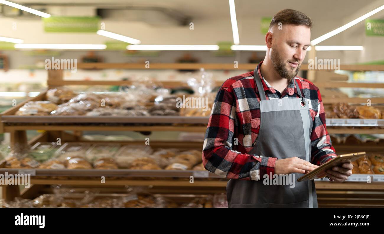 Confident supermarket clerk working with a digital tablet. Young handsome supervisor with tablet pc in the mall, background of bread shelves Stock Photo