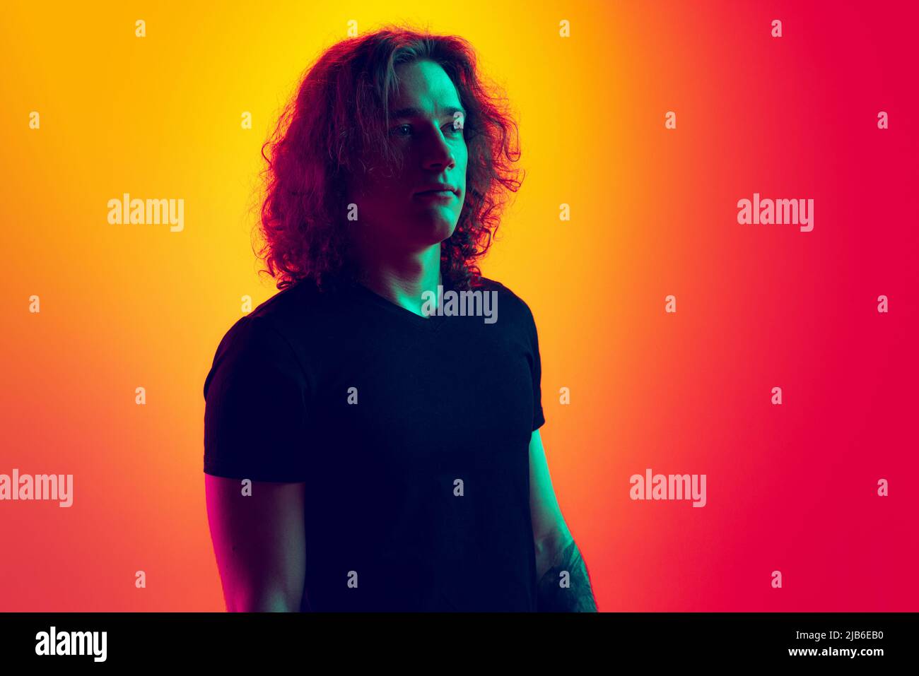 Young serious man with long curly hair posing isolated on gradient yellow-magenta background in neon. Concept of beauty, fashion, youth culture and Stock Photo