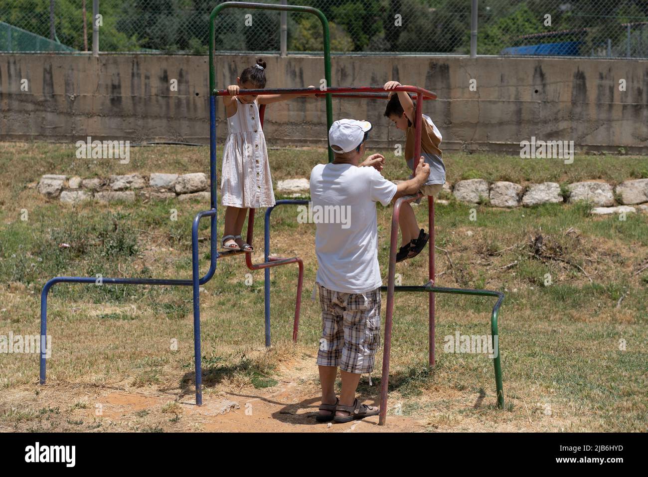 Grandchildren having fun with grandfather during sunny day in the playground. Back view senior man supporting her little grandson while playing at the outdoor playground on a lovely sunny day. Stock Photo