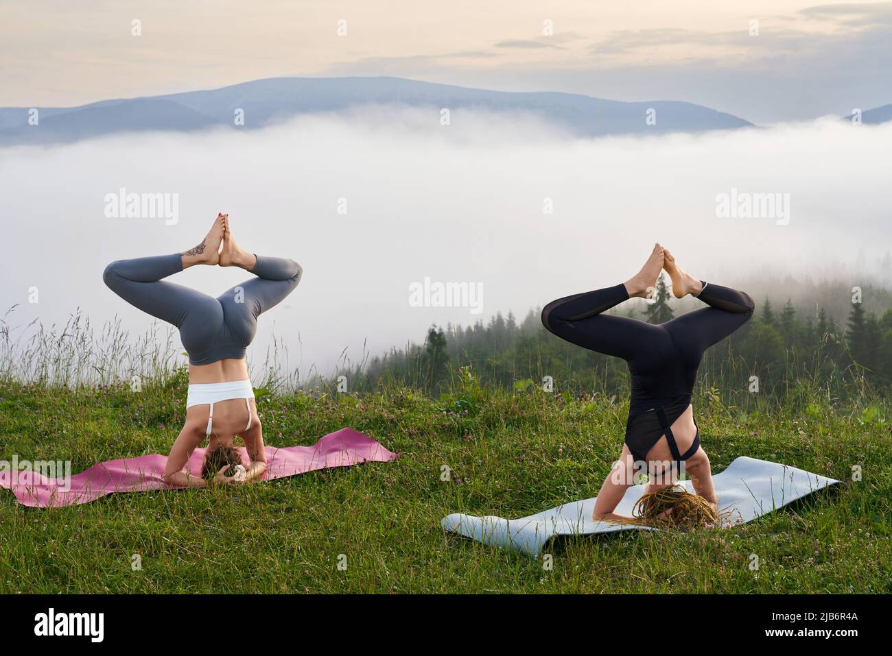 Two active women in sport clothes doing headstand exercises on yoga mat with amazing view of mountains on background. Concept of outdoors activity and balance. Stock Photo