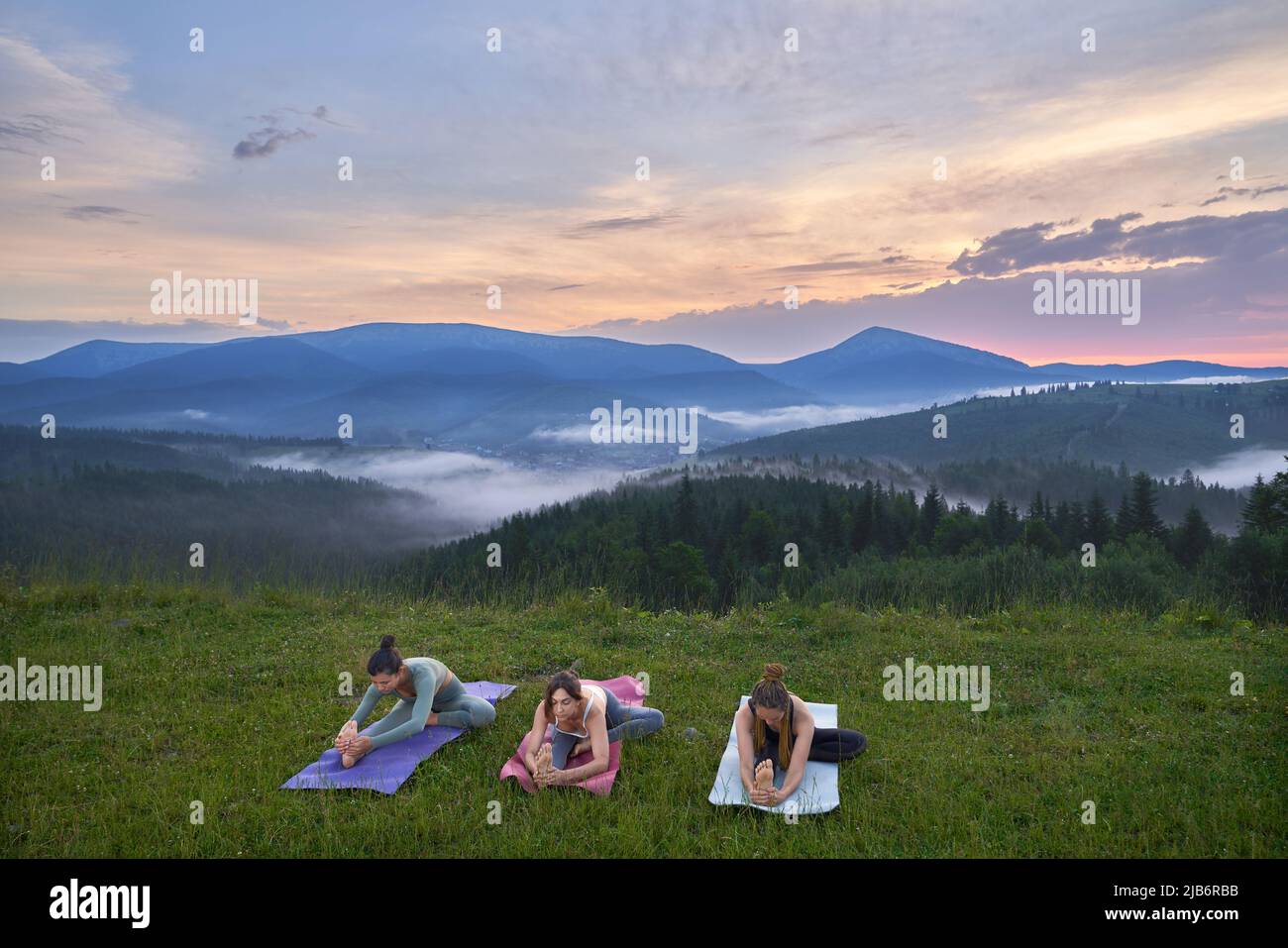 Three active women in sportswear stretching body on yoga mat with amazing sunset on background. Outdoors training for body and health care. Stock Photo