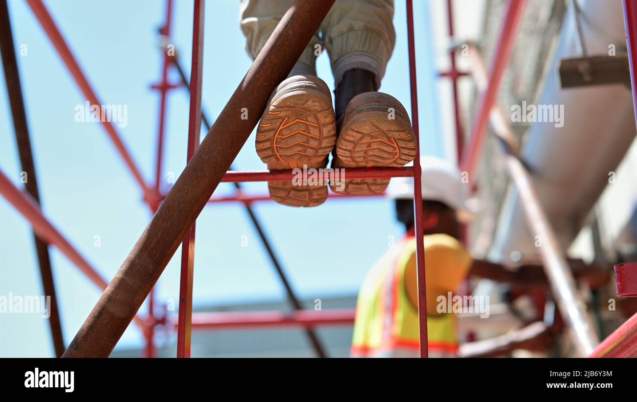 Two construction workers on scaffolding platform core drilling in sunshine. Steel toe capped work boot soles on red metal ladder rungs. Stock Photo