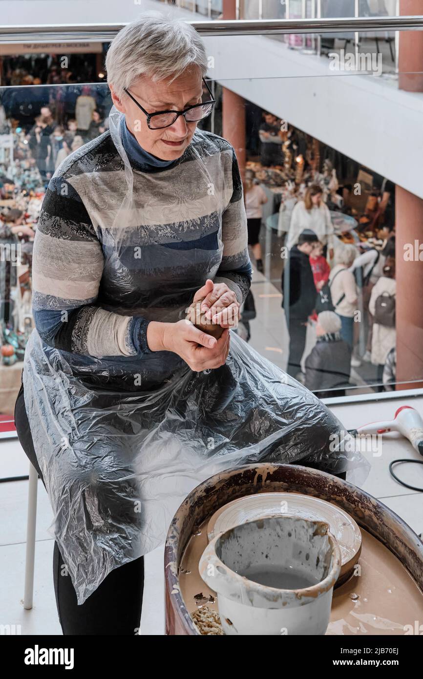 Senior gray-haired woman kneading clay in her hands to sculpt clay vase on potter's wheel. Stock Photo