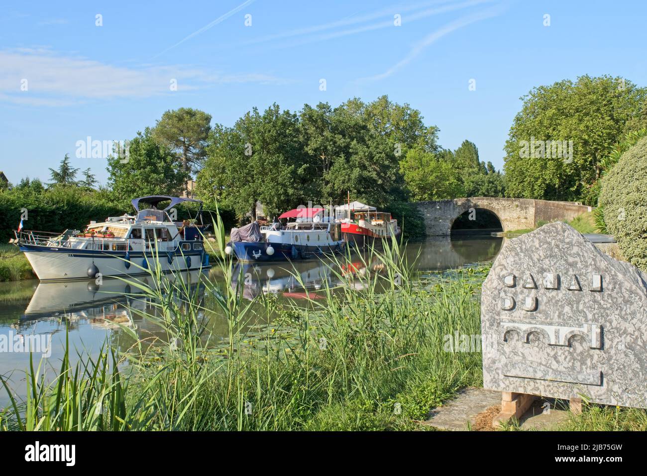 villesequelande Aude France 05.31.22 Canal du Midi summer landscape. Two cabin cruisers and a barge moored along a grass bank. Stone arched bridge. Stock Photo