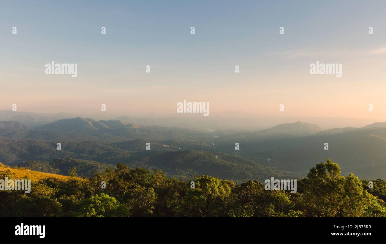 View across the Cardomom hills with view of rolling, forested landscape and trees under misty, clear sky at sunset near Thekkady, Kerala, India. Stock Photo
