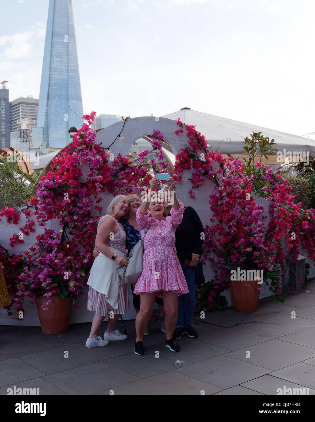 Women smiling and laughing as they take a selfie in front of artificial red and pink flowers at the Coppa Club with The Shard in the background. Stock Photo