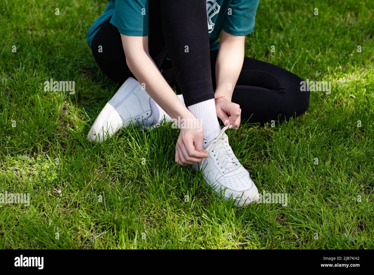 Close up view of young redhead woman wearing green tee standing on city park, outdoor tying lace running shoes getting ready for run. Jogging girl exe Stock Photo