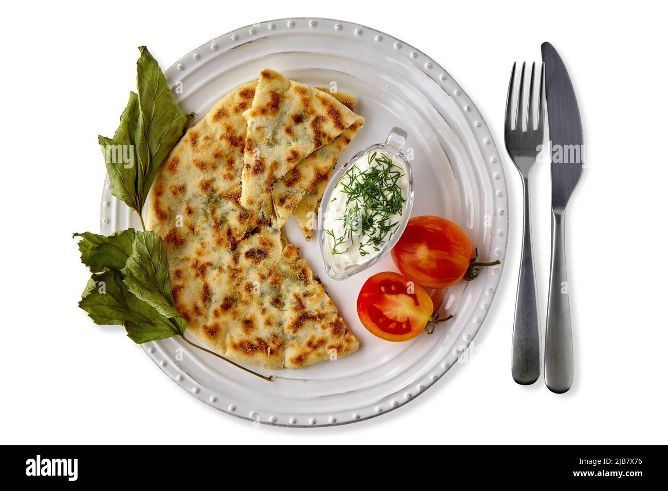 Flatbreads with cheese and herbs and yogurt sauce on a white plate with cutlery decorated with tomatoes and dried currant leaves. Vegetarian food Stock Photo