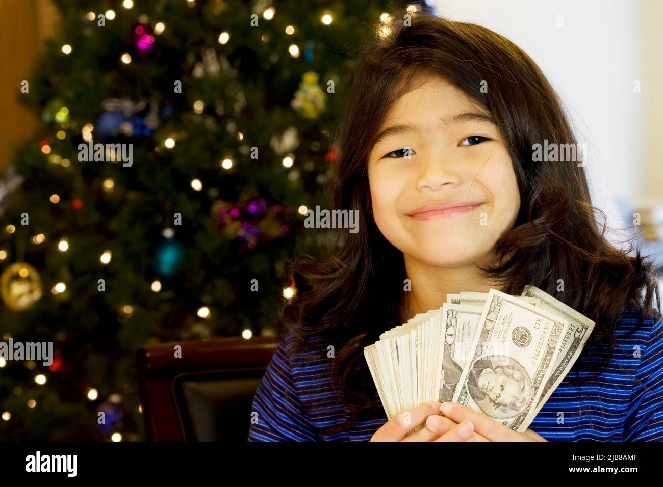 Little girl holding large amount of cash with Christmas tree in background Stock Photo