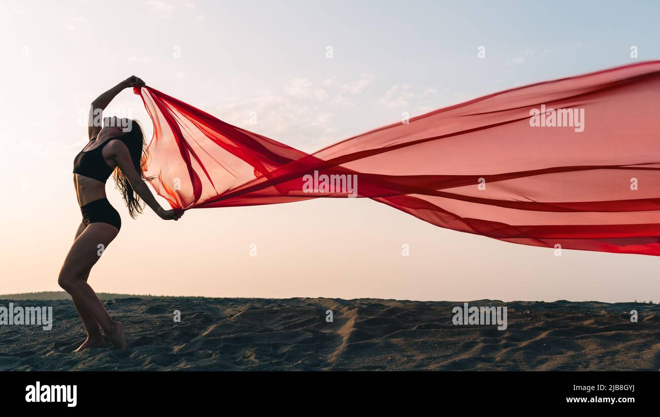 Elegant girl performs dance moves on the sand holding up long red fabric against sunset. Stock Photo