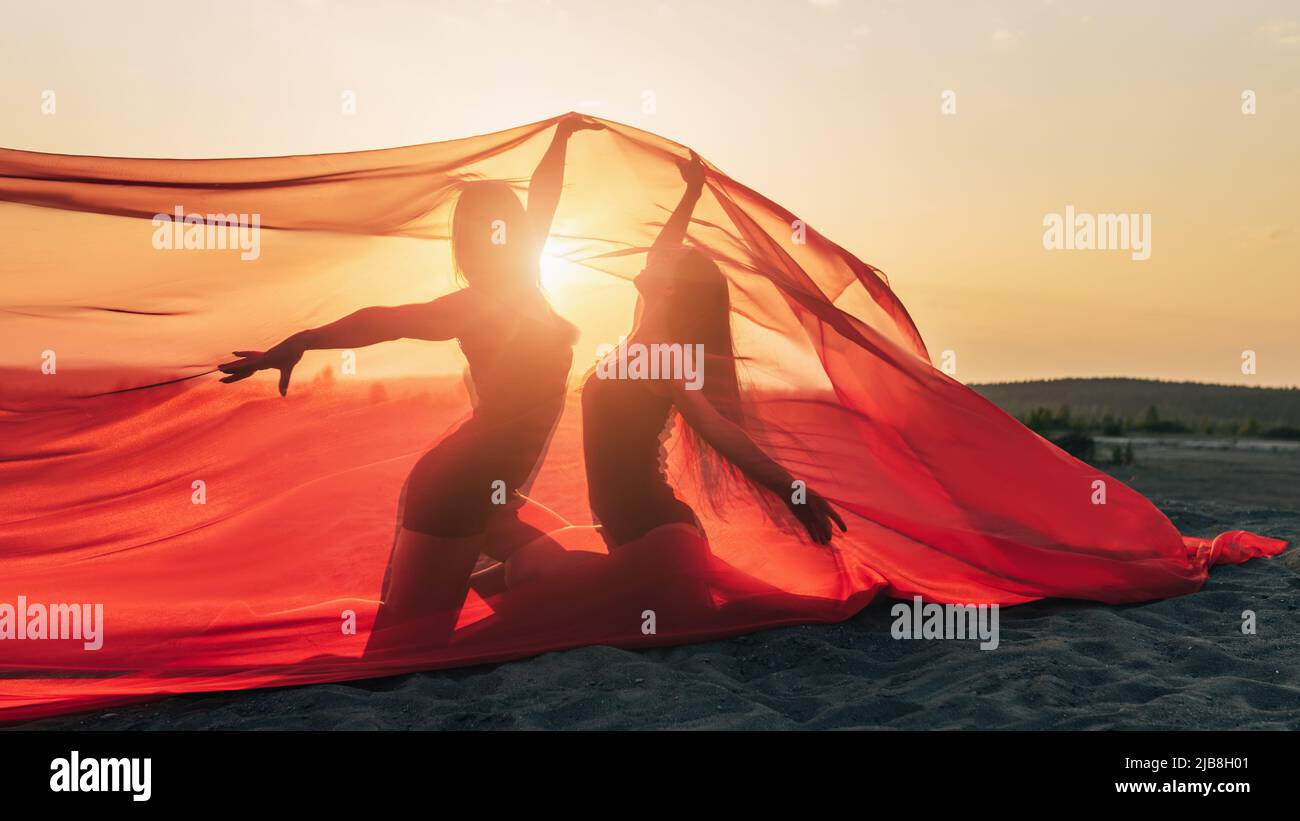 Elegant girl perform dance moves on sand holding up long red fabric against sunset. Stock Photo