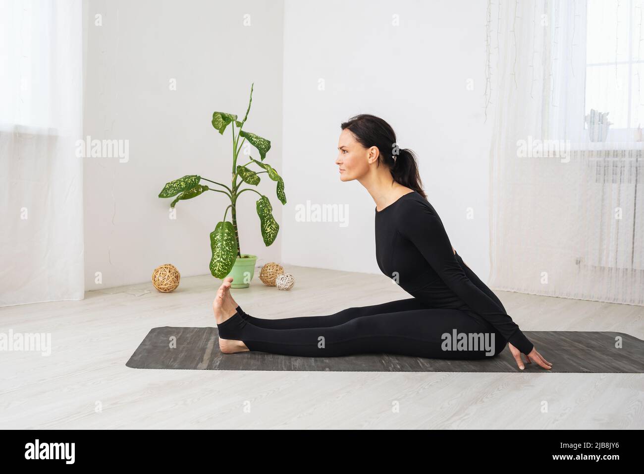 A woman in black sportswear does yoga, performs a forward bend, trains in a room, sitting on a mat Stock Photo