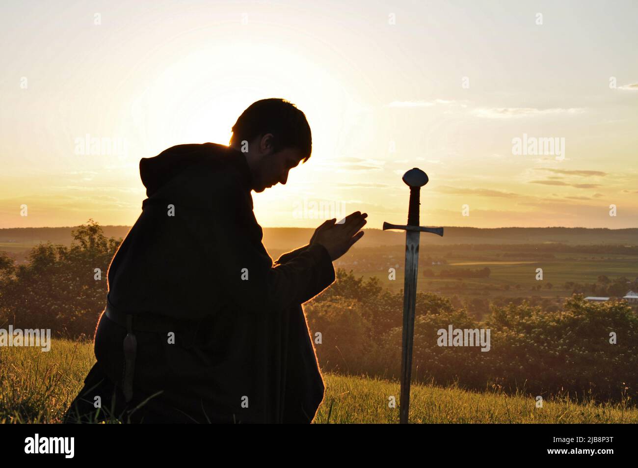 Knight praying silhouette and symbol of chivalry of a european knight, praying and kneeling in front of a sword in the sunset, with a burning sky. Stock Photo